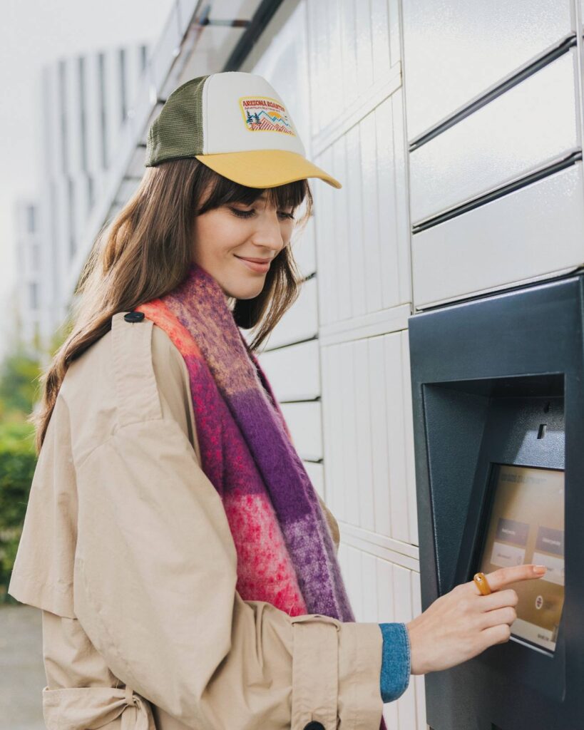 InPost / kobieta stoi przy paczkomacie. Ubrana jest w czapkę, jesienny płaszcz oraz wełniany, kolorowy szalik