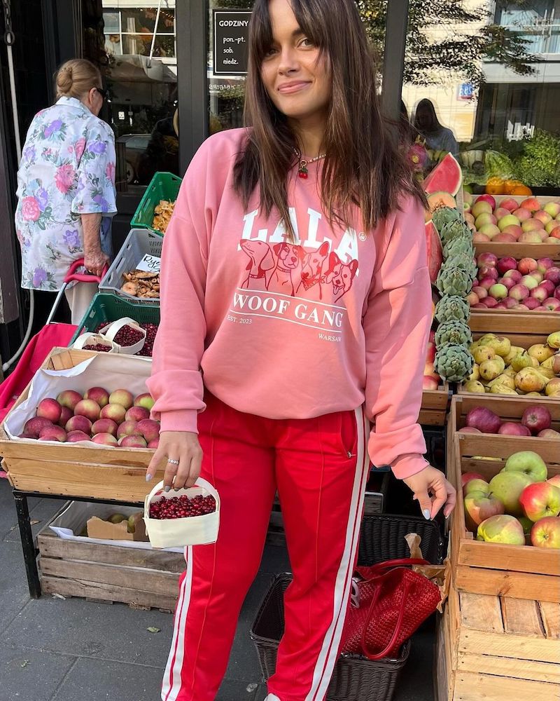 Elisa Minetti, właścicielka PLNY Lala. Na zdjęciu brunetka z długimi włosami w różowej bluzie z nadrukiem i czerwonych spodniach z białymi lampasami, na tle warzywniaka, trzyma w ręce koszyk z wiśniami.