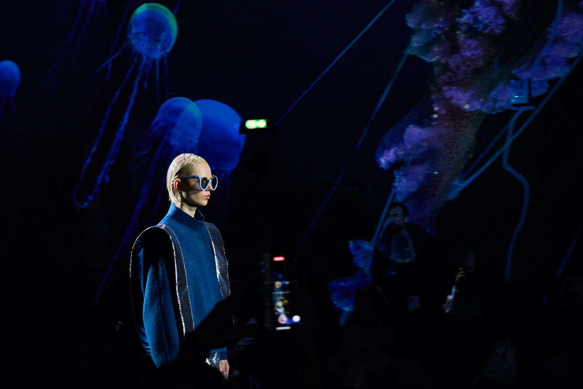 Francuska firma modowa Pierre Cardin i jej główny partner licencjonobiorca, firma Ahlers, zostały ukarane grywną przez Unię Europejską za nieuczciwe praktyki na rynku. Kara wynosi 5,7 mln euro. Pierre Cardin grzywna za złamanie zasad konkurencyjności. Na zdjęciu pokaz marki Pierre Cardin na sezon zima 2024. Model z blond włosami idzie po wybiegu w granatowej bluzce z golfem i w ciemnych okularach. Oprawa pokazu jest w kolorach ciemnego granatu.