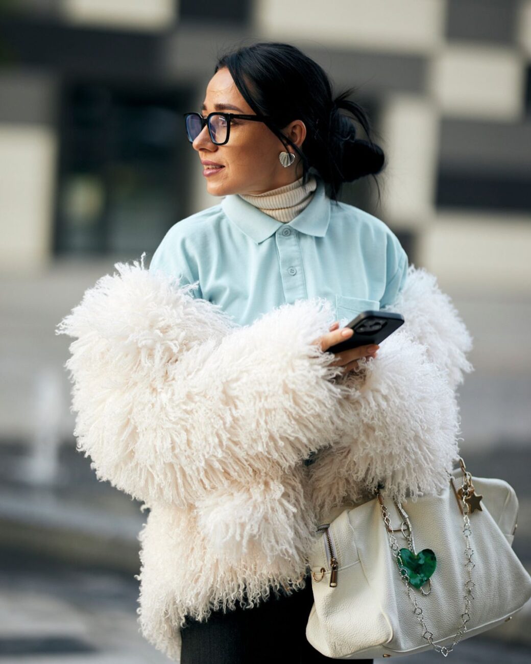 Obecność marki modowej w mediach nie jest już wyborem, a koniecznością. Konsumenci coraz częściej podejmują decyzje zakupowe pod wpływem treści, które pochłaniają scrollując ekrany smartfonów. Dominują dwie aplikacje: TikTok i Instagram. Gdzie publikować? Platformy oferują różne możliwości, a tym samym wymagają odmiennej strategii. Należy wziąć pod uwagę dotarcie do grupy docelowych, a także charakter marki, cele wizerunkowe i sprzedażowe. Sprawdzamy, gdzie najlepiej tworzyć content. Kobieta w niebieskiej koszuli i białym golfie, z futerkową, białą kurtką zarzuconą na ramię. Trzyma w ręce smartfona, a w drugiej białą torebkę z brelokami. Ma ciemne włosy związane w kok, okulary na nosie ora kolczyki w kształcie serduszek w uszach. Stoi na tle rozmytego budynku i patrzy w bok.