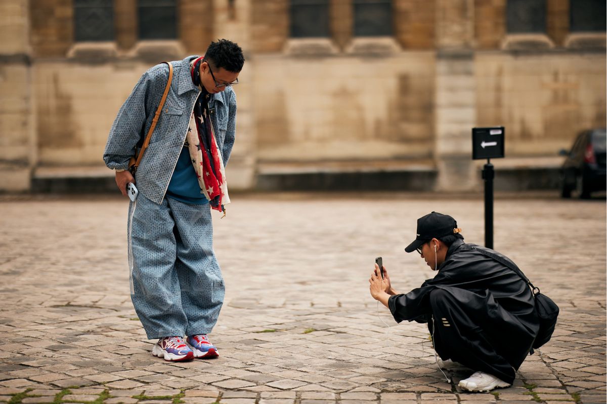 Obecność marki modowej w mediach nie jest już wyborem, a koniecznością. Konsumenci coraz częściej podejmują decyzje zakupowe pod wpływem treści, które pochłaniają scrollując ekrany smartfonów. Dominują dwie aplikacje: TikTok i Instagram. Gdzie publikować? Platformy oferują różne możliwości, a tym samym wymagają odmiennej strategii. Należy wziąć pod uwagę dotarcie do grupy docelowych, a także charakter marki, cele wizerunkowe i sprzedażowe. Sprawdzamy, gdzie najlepiej tworzyć content. Dwóch mężczyzn nagrywa content do mediów społecznościowych za pomocą smartfona. Model jest ubrany w szerokie jeansy oraz jeansową katanę i kolorowe sneakersy. Drugi mężczyzna, operator, jest ubrany na czarno i kuca z telefonem, robiąc kadr na sneakersy modela. Obok operatora stoi rozłożona lampa doświetlająca.