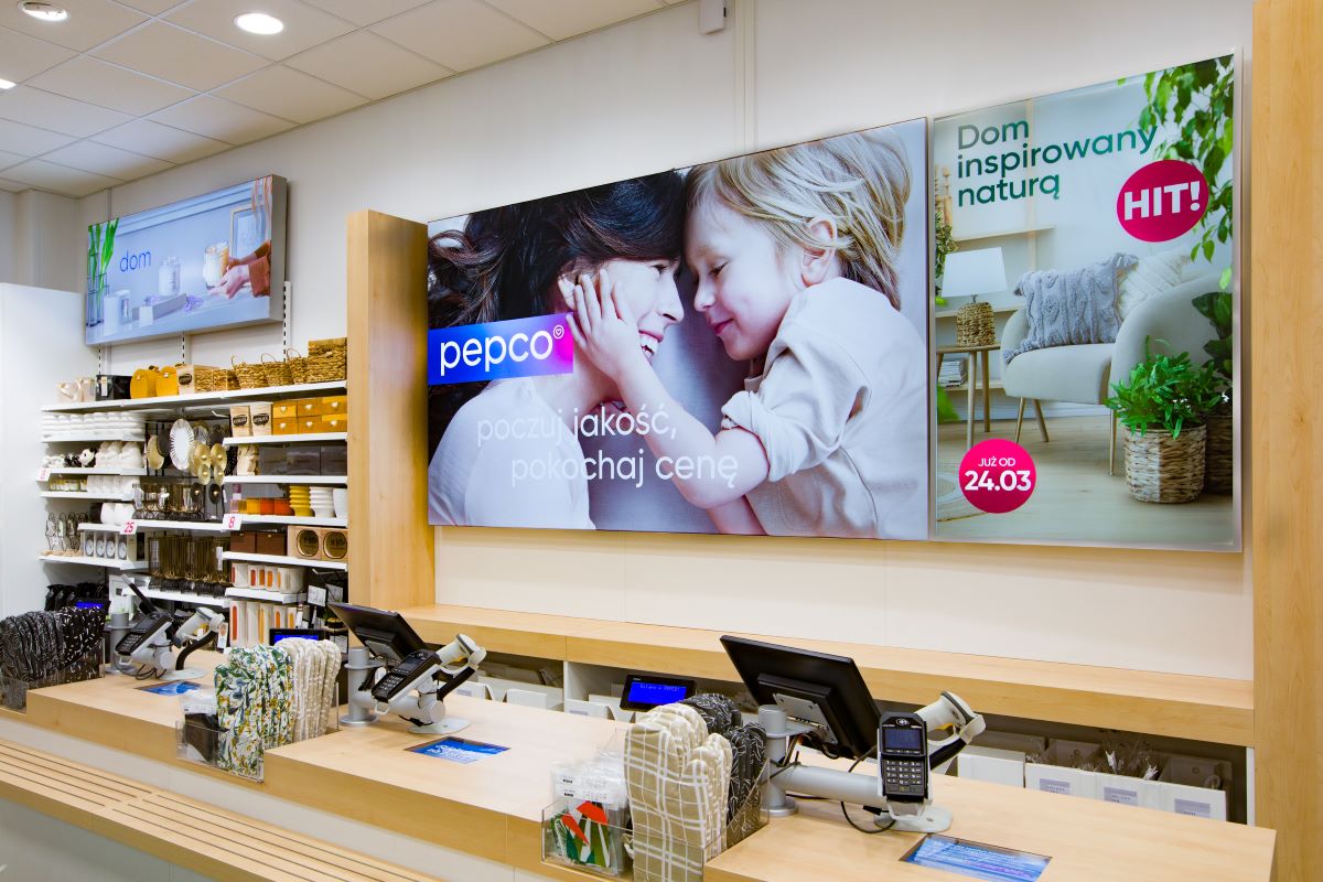 Ostatniego dnia września Grupa Pepco zamknęła rok obrotowy 2023/24. Opublikowany przez nią raport wskazuje na okres sumiennego rozwoju zakończonego solidnymi wzrostami. W przytoczonym terminie nie tylko jej zysk EBITDA rósł dwucyfrowo, również przychody przy kursach rzeczywistych wypracowane przez brytyjski podmiot urosły względem zeszłego roku o ponad 10 proc. Przeanalizowaliśmy wyniki finansowe Pepco oraz sprawdziliśmy, jakie są prognozy dotyczące dalszego rozwoju biznesu. Kasy w salonie stacjonarnym sieci sklepów Pepco. W tle reklamy produktów. W lewym sektorze zdjęcia znajdują się półki z ozdobami do wnętrz.