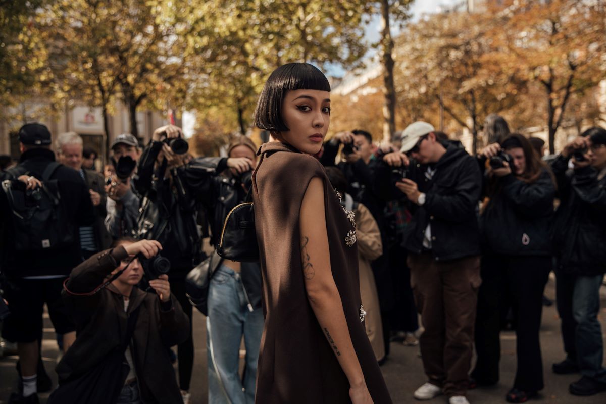 Stylowa kobieta z czarną przestępczością i wyrazistym makijażystem pozuje na tle grupy fotografów. Ma na sobie elegancką, ciemnobrązową kreację z ozdobnymi detalami. Scena, rozgrywająca się wśród jesiennych drzew, przypomina organizację najciekawszych wywiadów 2024