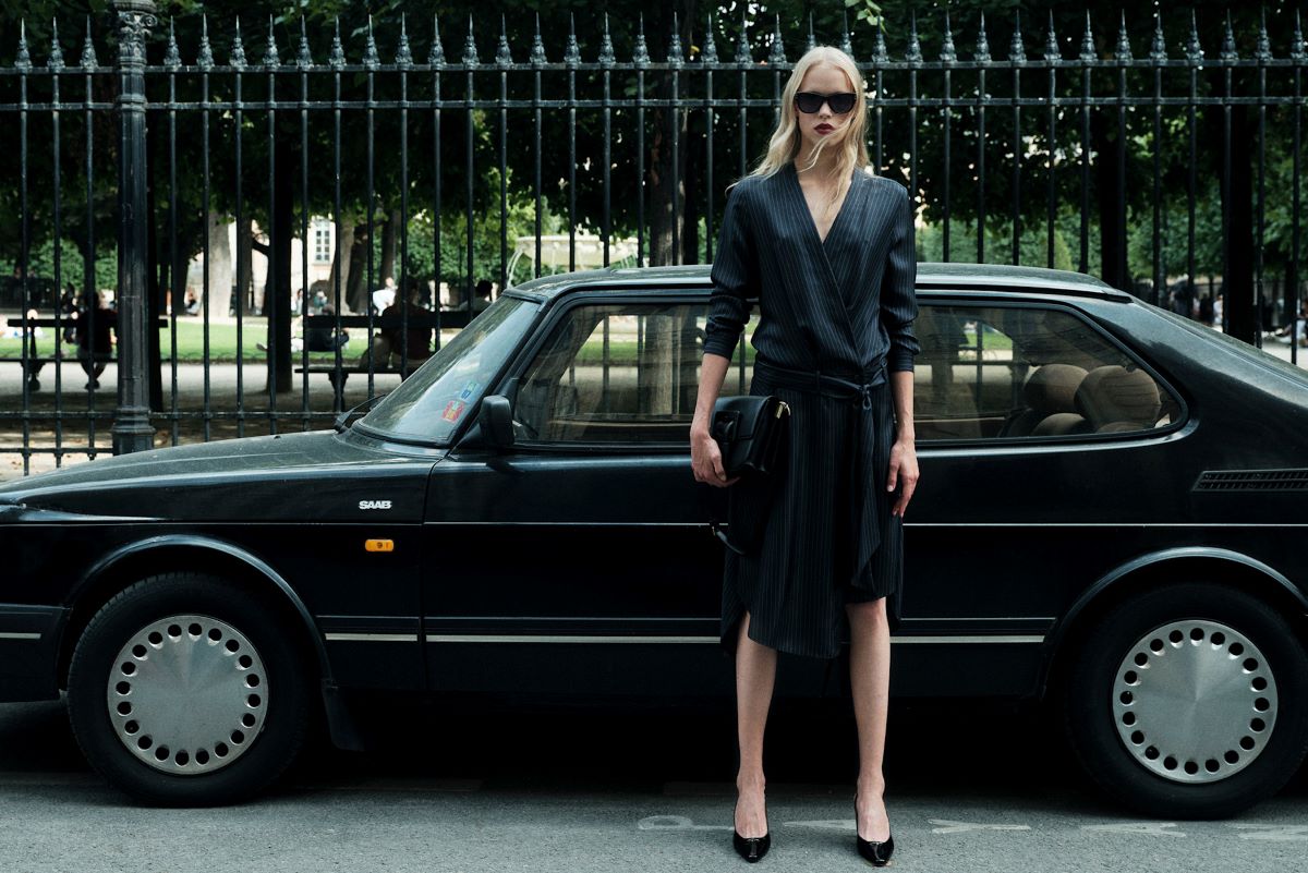 Kryzys dóbr luksusowych zmienia rynek, determinując wzrosty alternatywnych kategorii. Podróbki, vintage i szybka moda stają się pierwszymi wyborami konsumentów, którzy przed kilkoma miesiącami kupowali niemal wyłącznie artykuły najdroższych brandów. Czy to chwilowy trend, czy początek trwałej zmiany w branży? Sprawdzamy, kto naprawdę wygrywa na osłabieniu prestiżowych marek i jak te zmiany kształtują nowe oblicze rynku mody. Stylowa kobieta w eleganckiej, czarnej sukience i dużych okularach przeciwsłonecznych stoi na tle klasycznego czarnego samochodu Saab. Jej minimalistyczny strój i akcesoria podkreślają subtelne odniesienia do luksusu, jednocześnie symbolizując zmieniające się preferencje konsumentów wobec kryzys dóbr luksusowych.