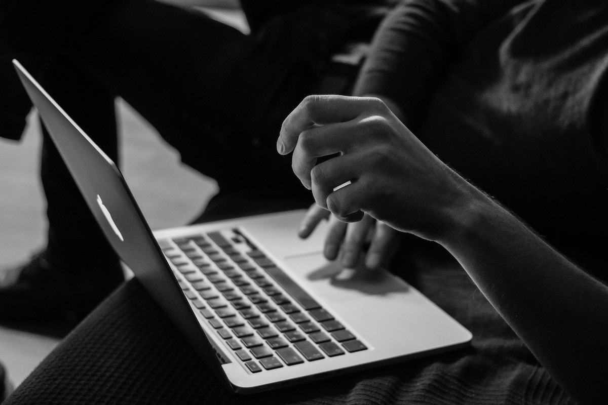 Na czarno-białej fotografii widać zbliżenie na osobę korzystającą z laptopa — w centrum kadru znajduje się ręka uniesiona nad klawiaturą, jakby w trakcie wykonywania gestu lub pisania. Laptop jest otwarty, a tło jest nieostre i ciemne, podkreślające sylwetkę użytkownika. Oszustwo na zwrotach pod Zgierzem.