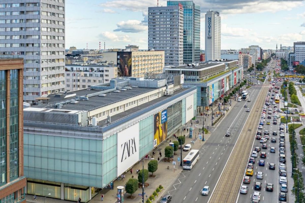 Widok z lotu ptaka na ulicę Marszałkowską w Warszawie, z dużym budynkiem handlowym, na którym znajduje się logo sklepu Zara na Marszałkowskiej. Po prawej stronie ulica z tramwajami i ruchem samochodowym, a w tle wysokie wieżowce, w tym hotel Novotel. Wzdłuż chodników poruszają się piesi.