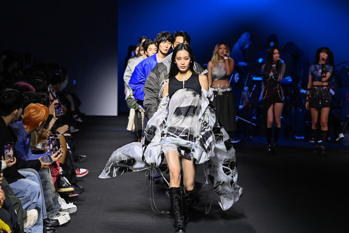 Przesyłki z Chin mogą wkrótce stać się droższe dla europejskich konsumentów. Wszystko za sprawą planowanego zniesienia limitu 150 EUR, który obecnie zwalnia je z opłat celnych. Branża e-commerce już rozpoczęła walkę o utrzymanie dotychczasowych zasad, wskazując, że nowe przepisy mogą negatywnie wpłynąć na rynek i portfele klientów. Jakie będą skutki zmian i czy Polska zdoła ochronić interesy konsumentów i przedsiębiorców? Na zdjęciu widzimy pokaz mody z grupą modeli i modelek prezentujących nowoczesne kreacje na wybiegu, w otoczeniu intensywnego oświetlenia i publiczności rejestrującej wydarzenie na smartfonach. W tle zauważalna jest scenografia oraz zespół muzyczny, co podkreśla dynamiczną atmosferę. Scena mogłaby symbolizować kreatywność i globalizację, podobnie jak przepływ towarów, takich jak przesyłki z Chin, które wpływają na różne branże, w tym modę.