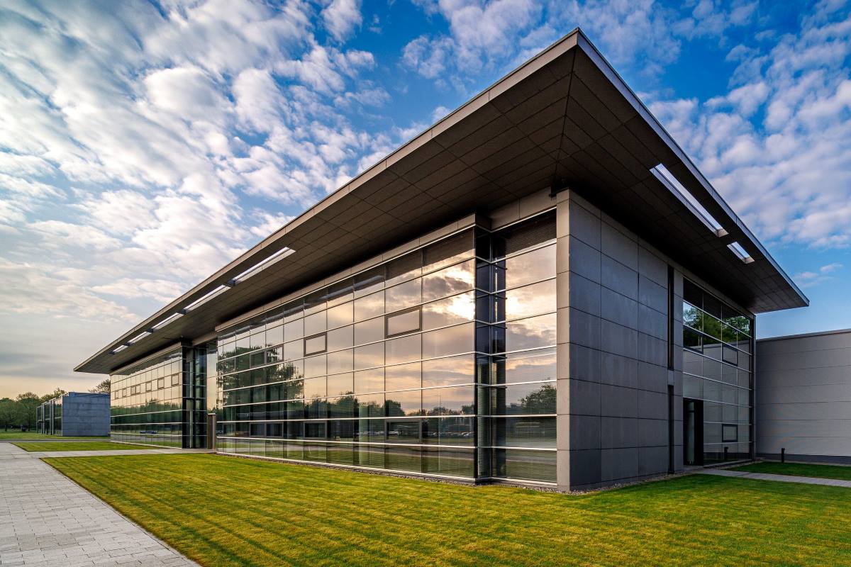 Spółka CCC zapowiedziała dużą inwestycję, która pozwoli wzmocnić jej pozycję na rynku i przyspieszyć rozwój marki HalfPrice. W Polkowicach powstanie nowoczesne centrum logistyczne, uzupełniające obecne zaplecze. Szacunkowa wartość całego projektu to około 200 mln zł. Jeszcze przed kilkunastoma miesiącami polski gigant borykał się z finansowymi turbulencjami, dzisiaj CCC inwestuje mocno myśląc o przyszłości i ustalaniu nowych standardów branżowych. Nowoczesny budynek z przeszkloną fasadą, otoczony zielonym trawnikiem i chodnikiem, na tle nieba z zachmurzeniem i prześwitującym słońcem. Zdjęcie przedstawia siedzibę Grupy CCC w Polkowicach, podkreślając rozwój i inwestycje firmy w infrastrukturę logistyczną.