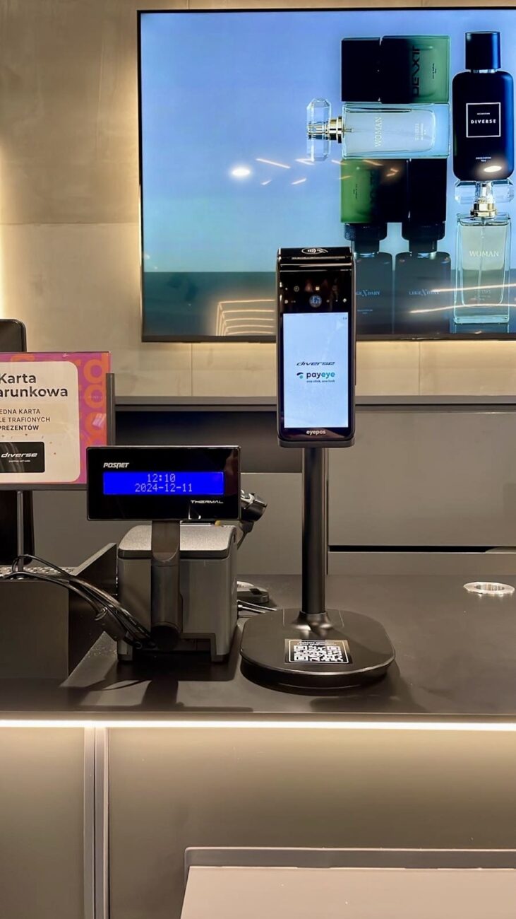 Diverse stawia na innowacje! Polska marka odzieżowa wprowadza do wybranych sklepów nowoczesne terminale płatnicze eyepos, stworzone przez firmę PayEye, które pozwolą na płatność jednym spojrzeniem. Dzięki tej współpracy klienci Diverse mogą korzystać z biometrycznych płatności, co znacząco podnosi komfort i szybkość obsługi. Zbliżenie na stanowisko kasowe w sklepie Diverse, wyposażone w nowoczesny terminal do płatności wzrokiem PayEye. Na ekranie w tle wyświetlana jest reklama perfum. Innowacyjne rozwiązania podkreślają nowoczesny charakter sklepu.