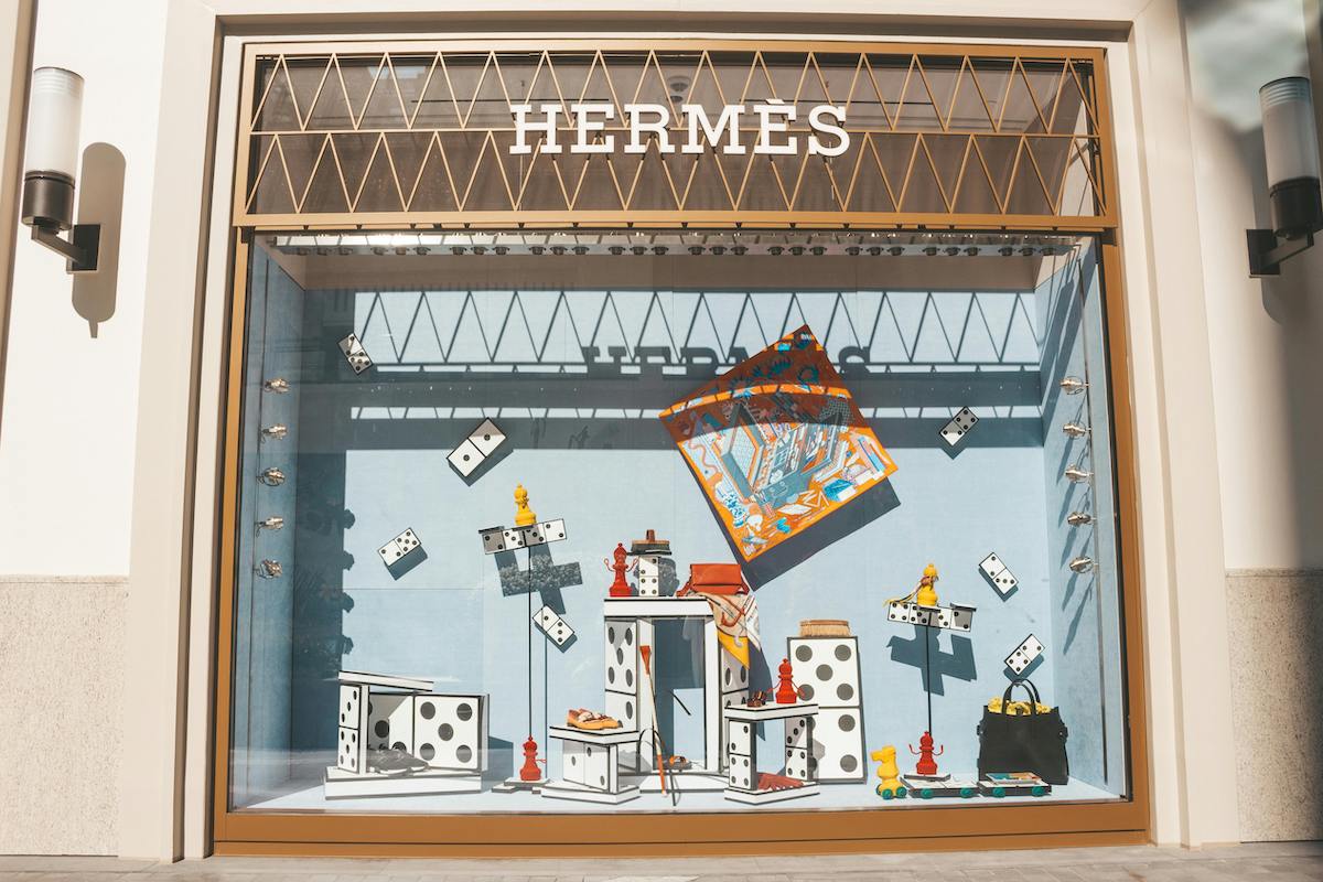 Wyniki finansowe Hermès za 2024 r. Witrynę sklepu Hermès zdobią duże domino, jaskrawe figury szachowe, pomarańczowo-niebieski szalik jedwabny pośrodku i czarna torba po prawej stronie; całość nawiązuje do tematyki gry planszowej.