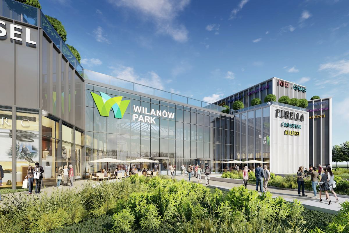 Wizualizacja nowego centrum handlowego Wilanów Park w Warszawie. Nowoczesny budynek ze szklaną fasadą i zielonymi tarasami. Na fasadzie widnieje logo „Wilanów Park” i nazwy sklepów. Przed wejściem znajduje się przestrzeń z roślinami, kawiarniami i spacerującymi ludźmi.