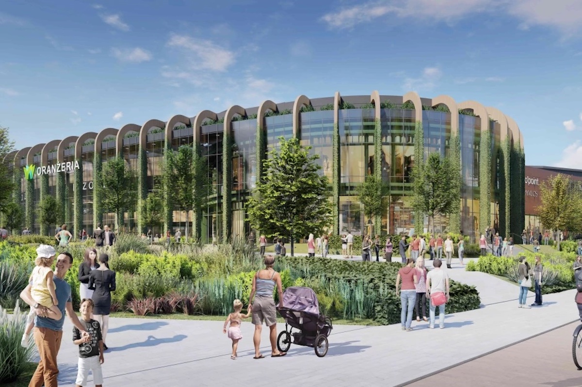 Wizualizacja nowoczesnego centrum handlowego Wilanów Park w Warszawie, z charakterystyczną zaokrągloną fasadą ozdobioną roślinami. Na budynku widnieje logo „Oranżeria”. Konstrukcja jest umieszczona w zielonej przestrzeni z drzewami i ścieżkami spacerowymi. Ludzie spacerują po ścieżkach, niektórzy z dziećmi i wózkami, co sprzyja przyjaznemu środowisku miejskiemu.
