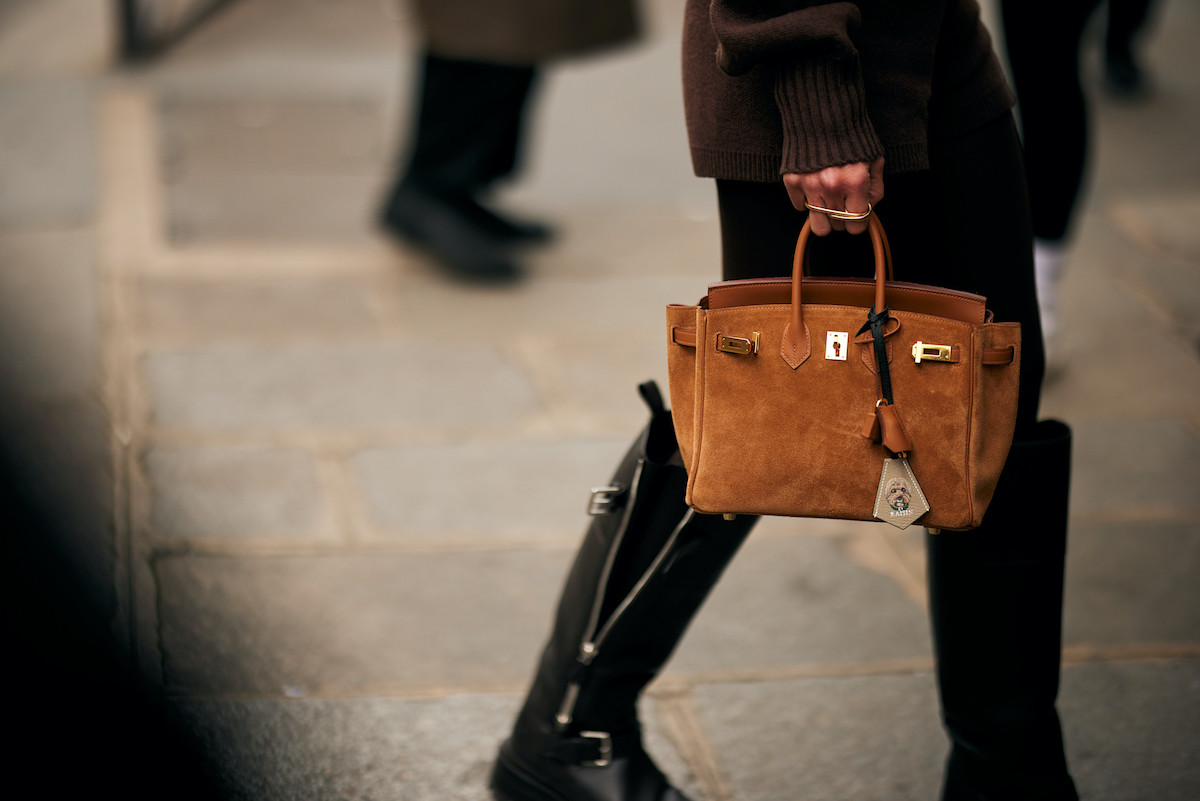 Hudson's Bay Najstarszy sprzedawca detaliczny w Kanadzie, na skraju bankructwa. Zbliżenie osoby idącej ulicą w czarnych skórzanych butach i brązowym swetrze, trzymającej elegancką brązową zamszową torebkę ze złotymi metalowymi zapięciami i zawieszką. W tle brukowany chodnik i rozmazane sylwetki przechodniów.