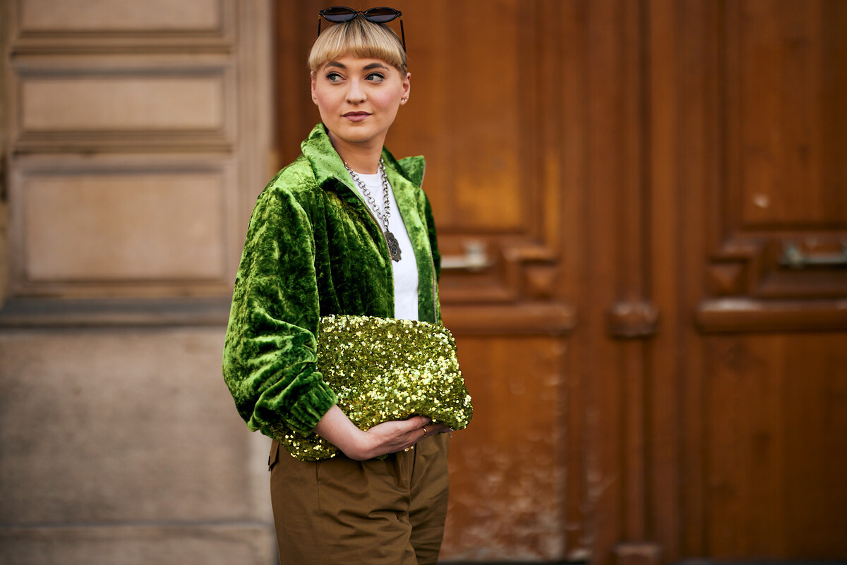 Forever 21. Młoda kobieta o jasnej cerze i blond włosach ułożonych w elegancki kok stoi przed drewnianymi drzwiami. Ma na sobie okulary przeciwsłoneczne. Jej strój składa się z obszernej zielonej aksamitnej bluzy, białego T-shirtu i beżowych spodni. Ma przy sobie dużą złotą i zieloną kopertówkę z cekinami. Jej szyję zdobi srebrny łańcuszek z dużym wisiorkiem. Spogląda w bok z delikatnym uśmiechem.