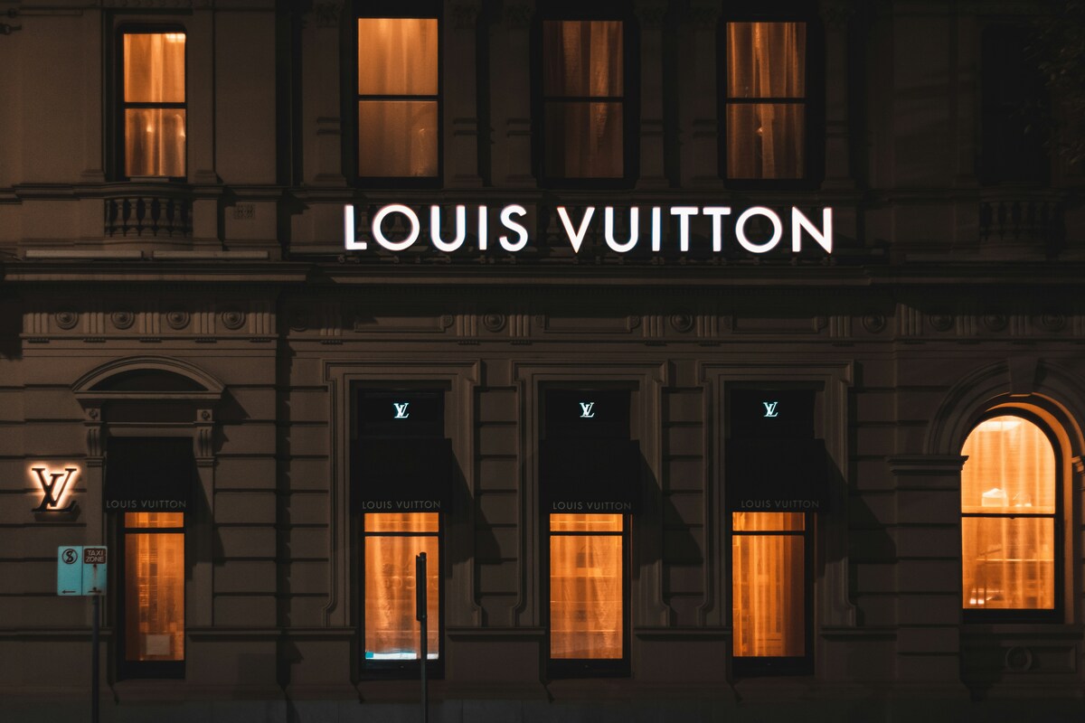 Zdjęcie sklepu Louis Vuitton zrobione nocą. Klasyczny budynek zdobi podświetlany na biało szyld z nazwą marki. Widoczne są również okna, oświetlone od wewnątrz.