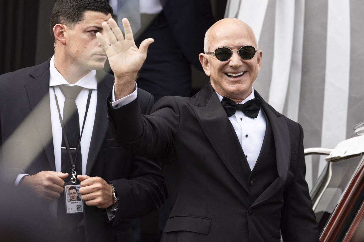 Założyciel Amazona, Jeff Bezos, macha i uśmiecha się w eleganckim czarnym garniturze, białej koszuli i czarnej muszce. Ma na sobie ciemne okulary przeciwsłoneczne, a za nim stoi ochroniarz w garniturze. Zdjęcie nawiązuje do sprzedaży akcji Amazona przez Jeffa Bezosa w 2025 roku.
