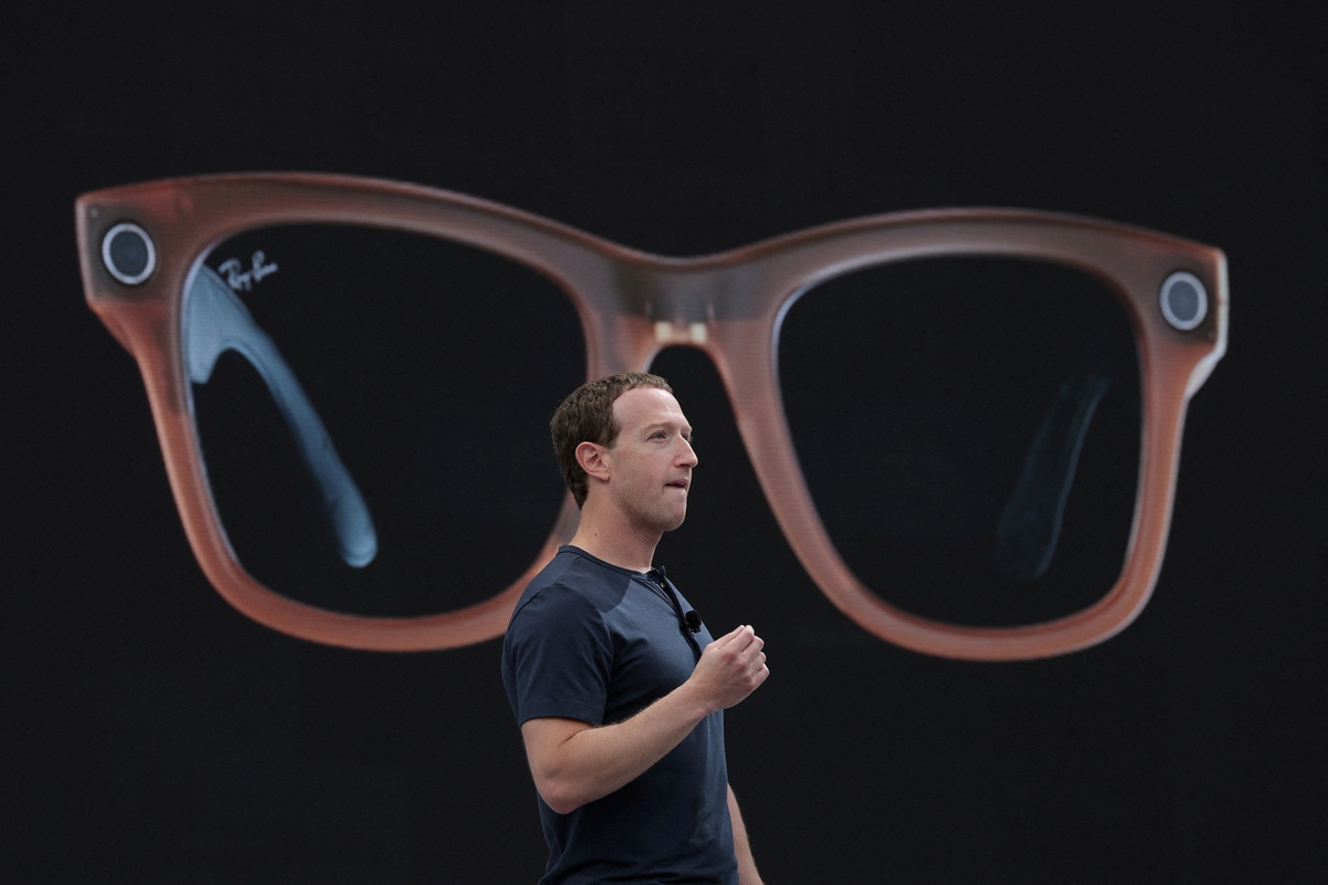 Zdjęcie Marka Zuckerberga w ciemnym T-shircie. Założyciel Facebooka prezentuje inteligentne okulary stworzone przez Meta i Ray-Ban. Inteligentne okulary wyświetlają się na ekranie za nim.