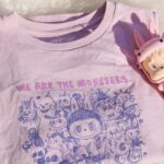 Labubu x UNIQLO. Zdjęcie koszulki z poprzedniej kolekcji stworzonej wspólnie przez Pop Mart i UNIQLO. Koszulka leży złożona na futrzanym dywanie, jest różowa i ozdobiona grafiką przypominającą rysunek piórem. Napisy są również różowe. Obok leży figurka Labubu z różowymi uszami.