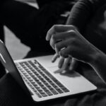 Czarno-białe zdjęcie przedstawia zbliżenie osoby korzystającej z laptopa – w centrum kadru widać dłoń uniesioną nad klawiaturą, jakby wykonywała gest lub pisała. Laptop jest otwarty, a tło jest rozmyte i ciemne, podkreślając sylwetkę użytkownika. Oszustwo związane ze zwrotem pieniędzy w okolicach Zgierza.
