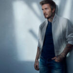 David Beckham, ubrany w jasnoszarą kurtkę typu bomber i granatowy T-shirt, opiera się o ścianę, prezentując nowoczesny, miejski styl BOSS.