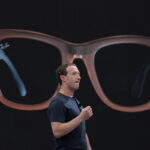 Zdjęcie Marka Zuckerberga w ciemnym T-shircie. Założyciel Facebooka prezentuje inteligentne okulary stworzone przez Meta i Ray-Ban. Inteligentne okulary wyświetlają się na ekranie za nim.