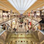 Czy Sadyba Best Mall zostanie zamknięta w 2026 roku? Zdjęcie przedstawia centrum handlowe Sadyba Best Mall, dwupiętrową konstrukcję, szerokie korytarze i liczne sklepy znanych marek, takich jak Mango. Widok z góry pokazuje schody ruchome prowadzące na niższy poziom, gdzie znajdują się kolejne sklepy i teren rekreacyjny z zielenią.