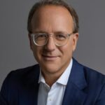 Prezes Estée Lauder. Zdjęcie portretowe. Elegancko ubrany mężczyzna w białej koszuli i granatowej marynarce, z lekko siwiejącymi włosami.