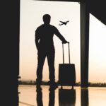 Limity płynów w bagażu podręcznym w 2025 roku. Co się zmienia? Na zdjęciu mężczyzna z walizką patrzy przez okno, przez które widać odlatujący samolot.