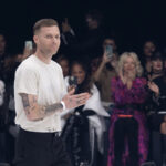 Projektant Matthew M. Williams po pokazie dla Givenchy. Ma wytatuowane ramiona i jest ubrany w biały T-shirt i czarne spodnie z paskiem. W tle widać owacje publiczności.