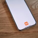 Telefon na drewnianym blacie. Biały wyświetlacz z pomarańczową ikoną Temu u dołu. Zdjęcie nawiązuje do zarzutów Komisji Europejskiej wobec Temu.
