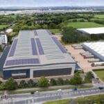 Zdjęcie przedstawia wizualizację nowoczesnego centrum logistycznego Panattoni, zlokalizowanego w Swindon w południowej Anglii. Obiekt szczyci się rozległym dachem pokrytym panelami słonecznymi, co podkreśla jego ekologiczny charakter. Na pierwszym planie widoczna jest droga prowadząca do centrum, a w tle – otaczająca zieleń oraz inne budynki przemysłowe i biurowe.
