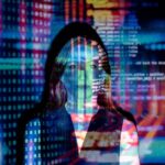 Agenci AI i agenci AI – czy zrewolucjonizują modę? Kobieta stoi w ciemnym otoczeniu, a na jej twarzy i ciele wyświetlane są cyfrowe obrazy przedstawiające kod komputerowy, liczby binarne i kolorowe linie reprezentujące dane. Obraz tworzy futurystyczną i technologiczną atmosferę, nawiązując do tematów związanych ze sztuczną inteligencją, cyberbezpieczeństwem lub przetwarzaniem danych.