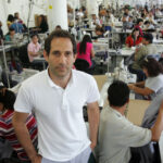 Dov Charney, założyciel American Apparel, stoi przed amerykańską szwalnią. Ma na sobie białą koszulkę polo, czarne włosy i ręce w kieszeniach. Zdjęcie nawiązuje do filmu dokumentalnego Netflixa o American Apparel: Total Disaster: The Rise and Fall of American Apparel.