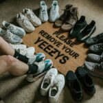 Zdjęcie przedstawia nogi mężczyzny siedzącego na dywanie. Wokół niego, w okręgu, ułożone są buty sportowe Adidas Yeezy w różnych kolorach i modelach, w tym Yeezy Boost 350 i Yeezy 700. W środku okręgu znajduje się okrągły dywan z napisem