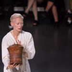 Tilda Swinton w Polsce – fryzura Daniela Gryszke. Na zdjęciu Tilda Swinton, z krótkimi, jasnymi włosami i luźnym, białym strojem, trzyma drewniane naczynie przypominające bęben lub pojemnik. Znajduje się na scenie lub w przestrzeni, w której odbywa się przedstawienie, a w tle widać siedzących widzów.