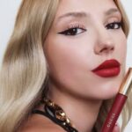 Coty – Sprzedaż marki. Zdjęcie modelki z rozpuszczonymi blond włosami, reklamującej czerwony błyszczyk lub szminkę. Kobieta ma na ustach czerwoną szminkę i makijaż w stylu „kocie oczy”.