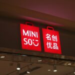 Sklep Miniso Polska. Zdjęcie szyldu z logo Miniso – czerwone torby zakupowe z białym napisem. Zawieszone nad wejściem do sklepu.