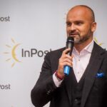 Rafał Brzoska, założyciel InPost, na konferencji. Ubrany w garnitur, stoi z mikrofonem w dłoni i przemawia. Za nim widnieje baner z logo InPost.