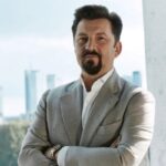 Portret mężczyzny w eleganckim szarym garniturze stojącego przed przeszkloną szybą z widokiem na nowoczesną panoramę miasta. Jego profesjonalny wygląd podkreśla zaangażowanie w ambitne projekty – CCC inwestuje w rozwój i przyszłość swojej sieci logistycznej oraz HalfPrice.
