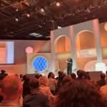 Scena ceremonii wręczenia nagród LVMH Innovation Award, elegancko udekorowana w ciepłych odcieniach pomarańczu i beżu, przedstawia kobietę w ciemnym garniturze, która prowadzi wydarzenie, a jej wizerunek jest również wyświetlany na dużym ekranie z boku sceny. Przed sceną zasiada liczna publiczność, a słowa…
