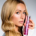 Paris Hilton reklamuje swoją nową markę kosmetyków do pielęgnacji skóry Parívie, trzymając w dłoni butelkę produktu.