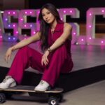 Lucy Guo, najmłodsza miliarderka na świecie, stoi przed neonowymi reklamami w jaskraworóżowym stroju i białych trampkach, trzymając deskorolkę.