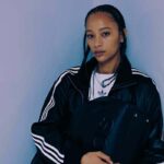 Adidas debiutuje ze swoją luksusową kolekcją A-Type. Na zdjęciu modelka ma na sobie jeden z nowych projektów marki – czarną bluzę z kultowymi trzema białymi paskami i biały T-shirt. Modelka trzyma czarną skórzaną torbę.