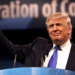USD/PLN Donald Trump pokazuje symbol zwycięstwa prawą ręką z podium. Ma na sobie białą koszulę, niebieski krawat i czarną marynarkę.