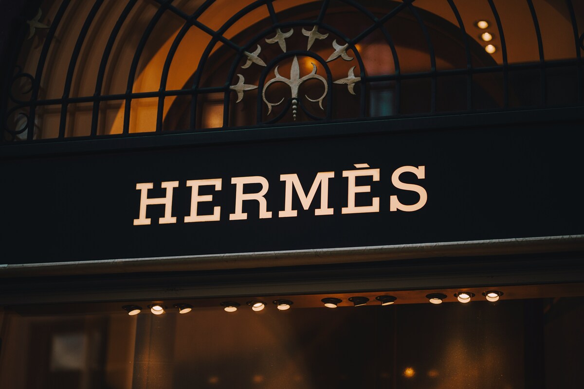 Wyniki finansowe Hermès. Zdjęcie podświetlanego szyldu, zrobione nocą. Jasne litery oznaczają Hermès. W tle widoczne są ozdobne kratki i elementy budynku.