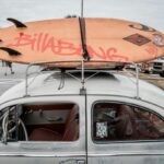 Quiksilver, Billabong i Volcom zamykają sklepy. Na zdjęciu widać jasny, klasyczny samochód z deską surfingową Billabong przymocowaną do bagażnika dachowego. W tle widać zaparkowane pojazdy i ludzi na parkingu, a także fragmenty drzew i budynków.