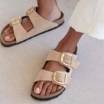 Zdjęcie podróbek butów Birkenstock w Indiach. Mocno opalona kobieta w długich białych spodniach ma na sobie beżowe klapki Birkenstock. Ma naturalne paznokcie.