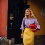 Ulta Beauty debiutuje w Europie. Kiedy pojawi się w Wielkiej Brytanii? Na zdjęciu uśmiechnięta blond modelka w fioletowym T-shircie i żółtej spódnicy, trzymająca czerwoną kopertówkę.