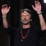 Pierpaolo Piccioli unosi ręce po pokazie. Włoski projektant ma na sobie czarną bluzkę, zegarek i czerwony męski wisiorek.