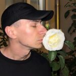 Zbliżenie twarzy założyciela Pinoko Models. Wojciech Tubaja, ubrany w czarny T-shirt i czarną czapkę baseballową, wącha białą różę w ogrodzie przed domem.
